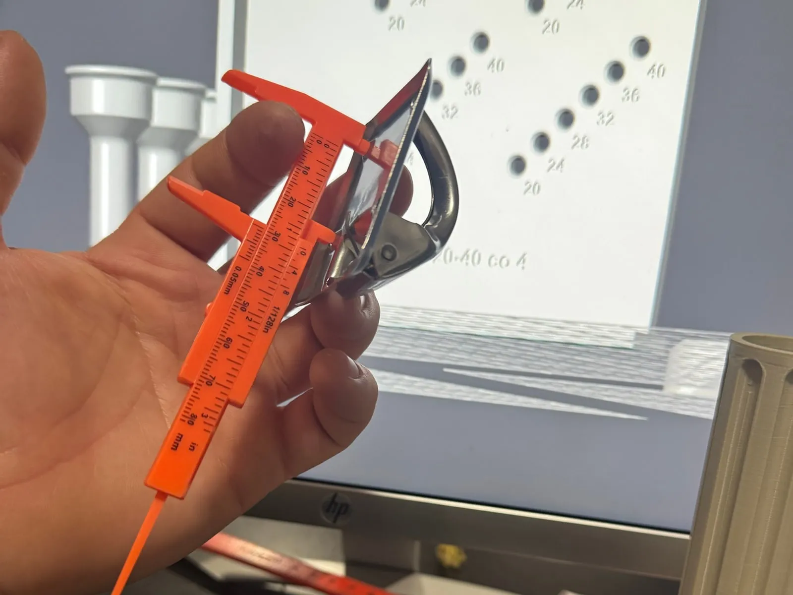 Dłoń z suwmiarką mierzy metalowy zatrzask; w tle model części w programie do druku 3D i wydrukowana rurka